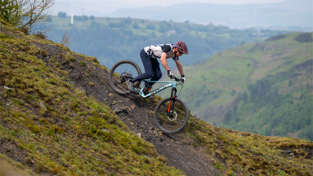 A mountain biker wearing a purple helmet navigates a steep dirt trail on a full-suspension bike with a light blue frame, set against a backdrop of green rolling hills.
