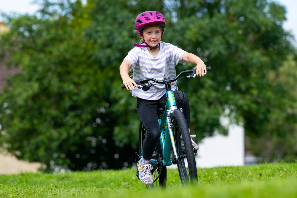 A young girl riding a teal mountain bike on a grassy lawn, wearing a pink helmet, a white and gray striped t-shirt, and black pants, with a blurred green tree background.