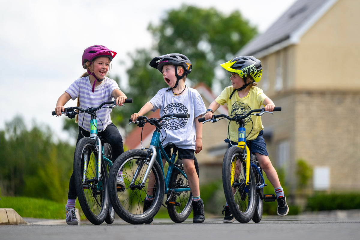 Three children wearing helmets ride colorful children's bicycles on a paved path outdoors, with a blurred house and greenery in the background.