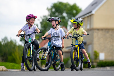Three children wearing helmets ride colorful children's bicycles on a paved path outdoors, with a blurred house and greenery in the background.