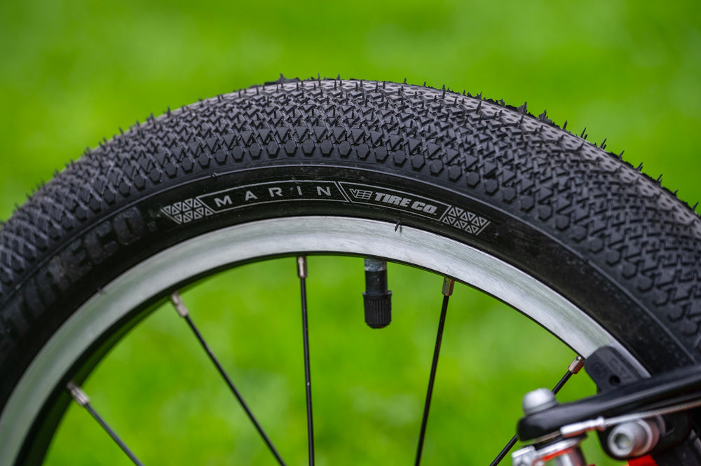 Close-up of a black bicycle tire with visible tread and sidewall text reading 'MARIN TIRE CO' and 'MICHELIN', mounted on a wheel with a silver rim against a blurred green grass background.
