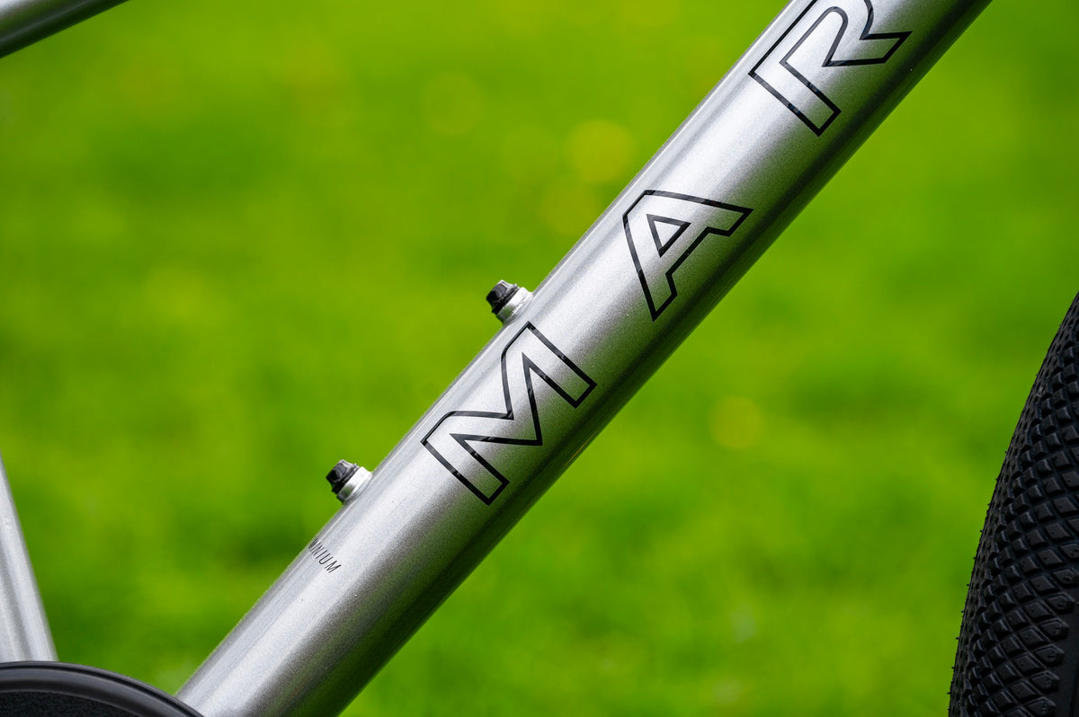 Close-up of a silver metallic bicycle frame tube with the brand name 'MAR' printed in black outline, set against a blurred bright green background.