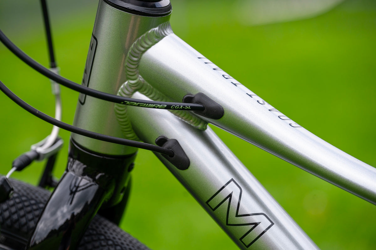 Close-up of a silver mountain bike frame with black cables routed through black plastic loops, set against a blurred green outdoor background.