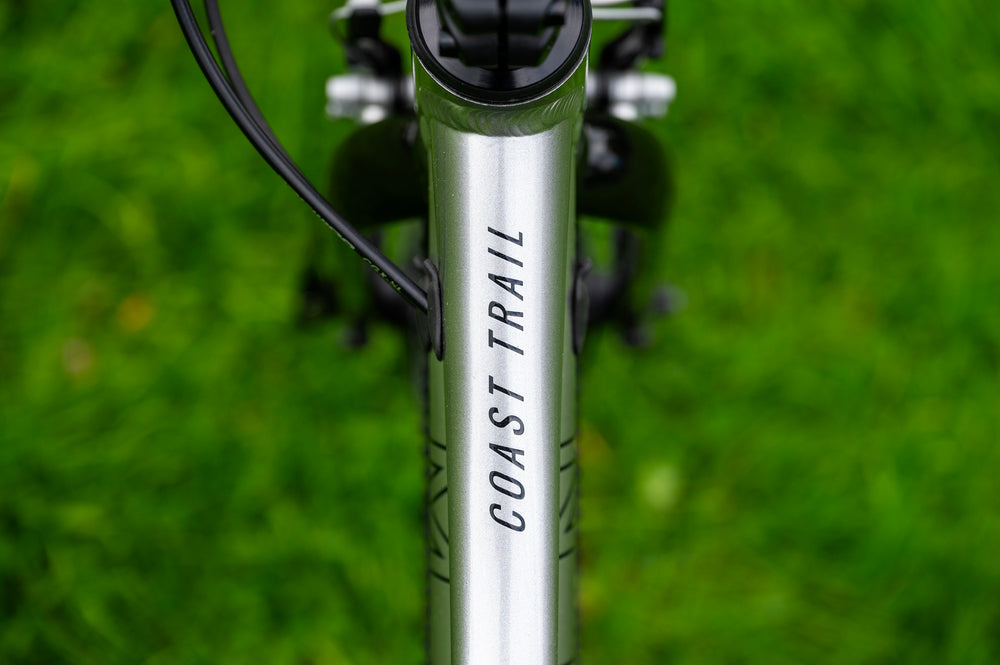 Close-up of a silver bicycle frame with 'COAST TRAIL' printed vertically in black, set against a blurred green grass background.