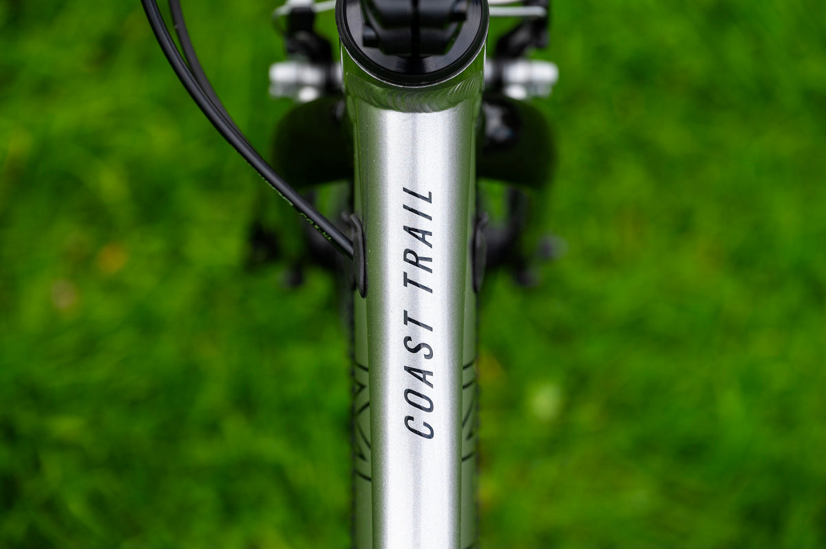 Close-up of a silver bicycle frame with 'COAST TRAIL' printed vertically in black, set against a blurred green grass background.