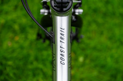 Close-up of a silver bicycle frame with 'COAST TRAIL' printed vertically in black, set against a blurred green grass background.