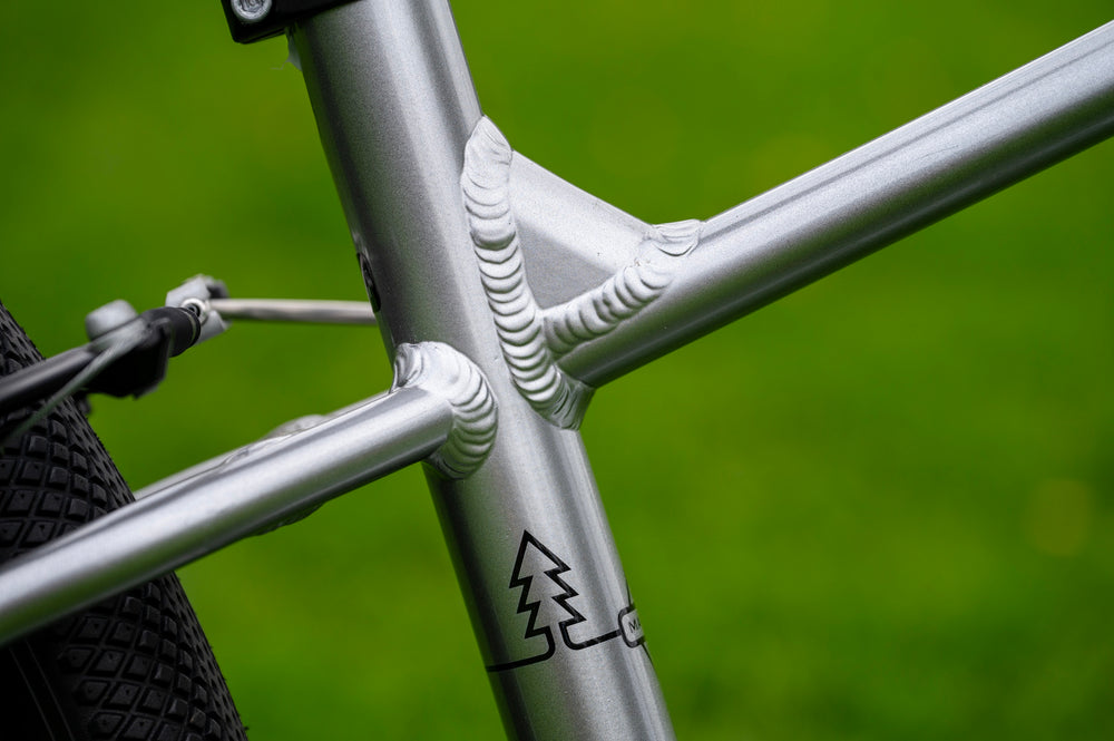 Close-up of a silver aluminum bicycle frame with a textured weld joint and a black tree logo, set against a blurred green outdoor background.