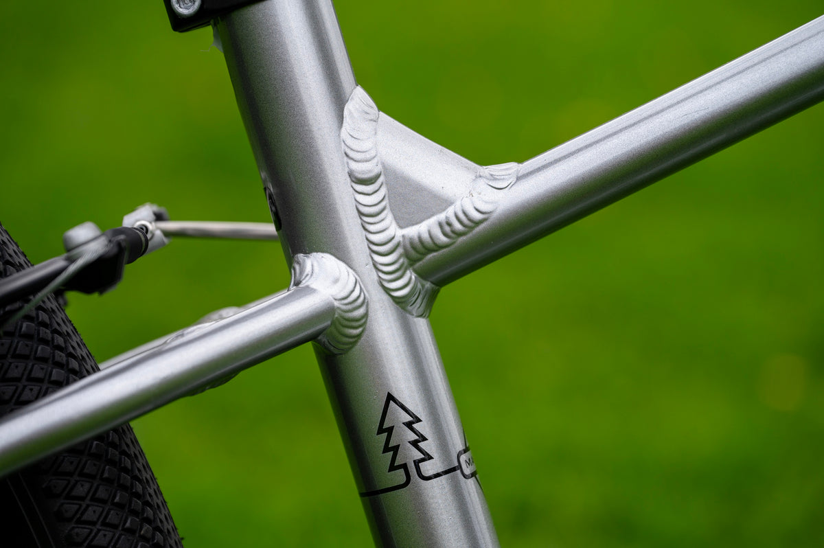 Close-up of a silver aluminum bicycle frame with a textured weld joint and a black tree logo, set against a blurred green outdoor background.