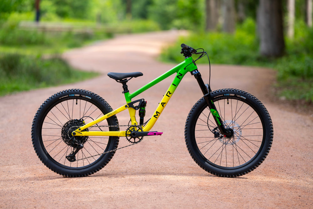 A mountain bike with a bright green and yellow frame, black tires, and a black saddle, parked on a dirt path in a lush green outdoor setting.