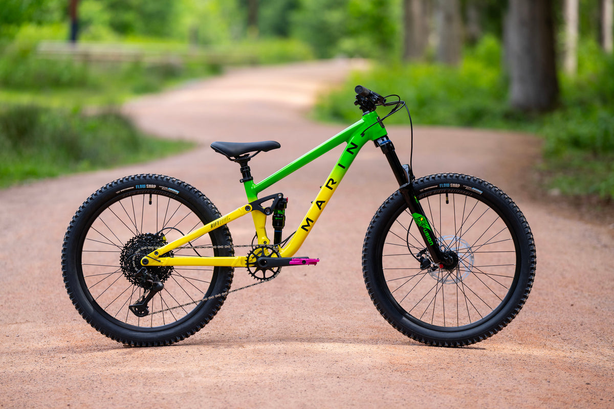 A mountain bike with a bright green and yellow frame, black tires, and a black saddle, parked on a dirt path in a lush green outdoor setting.