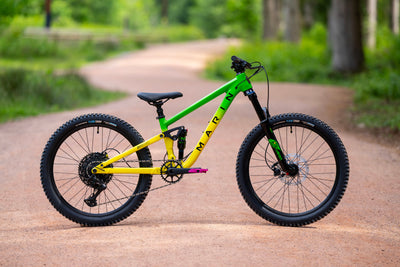 A mountain bike with a bright green and yellow frame, black tires, and a black saddle, parked on a dirt path in a lush green outdoor setting.