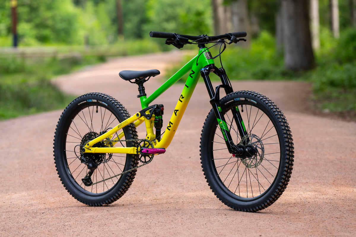A Marin full-suspension mountain bike with a bright lime green and yellow frame, parked on a dirt path in a forest setting.