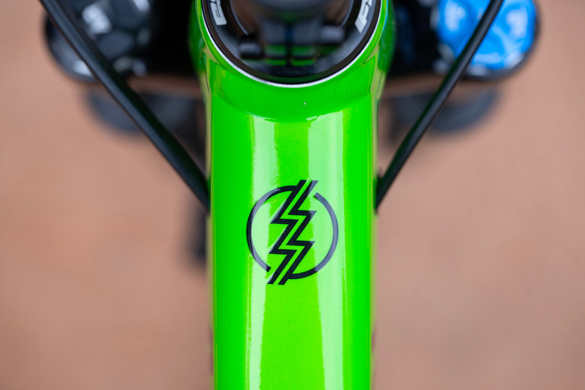 Close-up of a bright green bicycle frame with a black lightning bolt logo, showing part of the handlebar and blurred background.