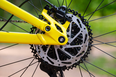 Close-up of a bicycle's rear wheel hub, showing a bright yellow frame, a silver disc brake rotor, a black brake caliper with 'TORKPRO' branding, and a black cassette on a blurred green outdoor background.