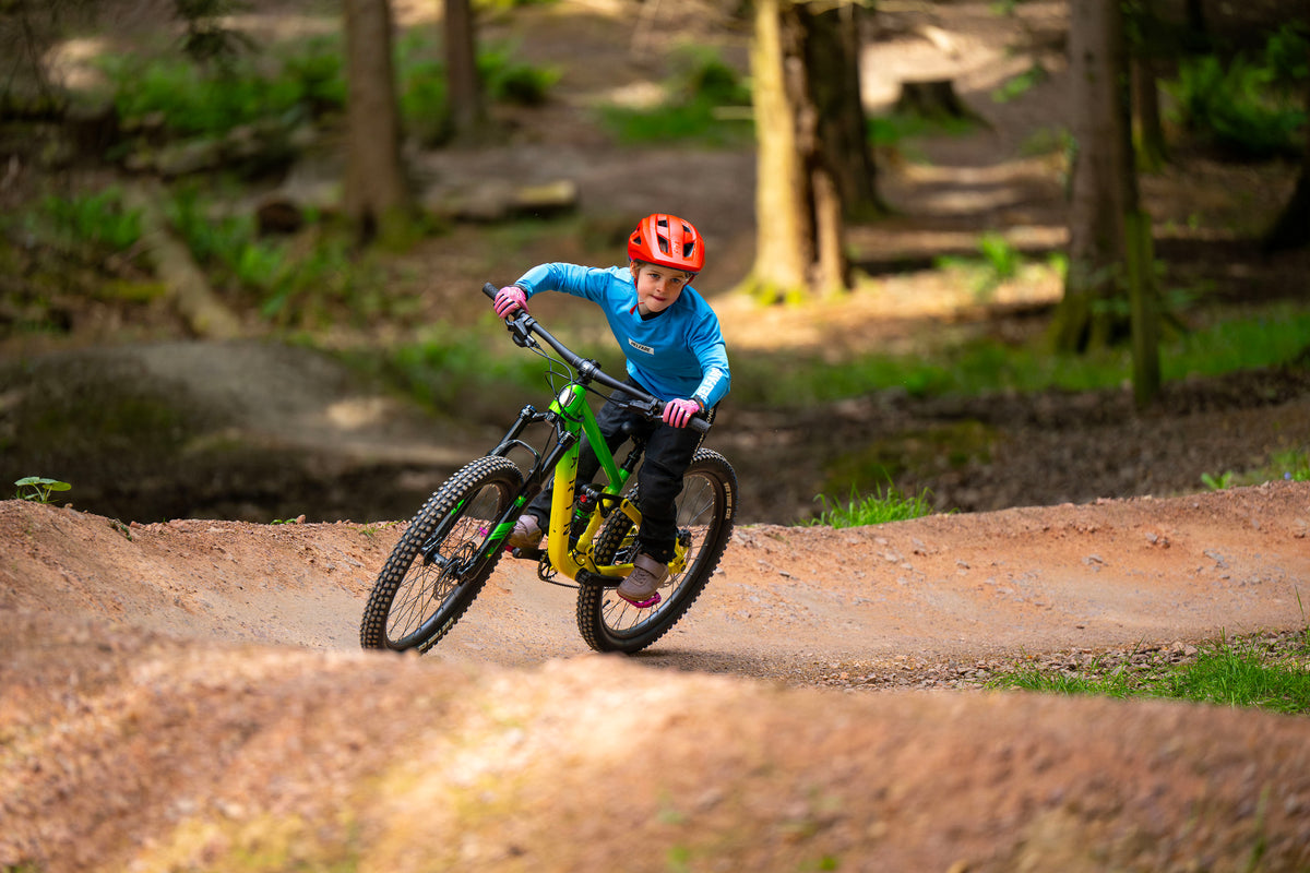 A child wearing a red helmet and blue jacket rides a green and yellow mountain bike on a dirt trail in a forest.
