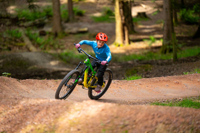 A child wearing a red helmet and blue jacket rides a green and yellow mountain bike on a dirt trail in a forest.