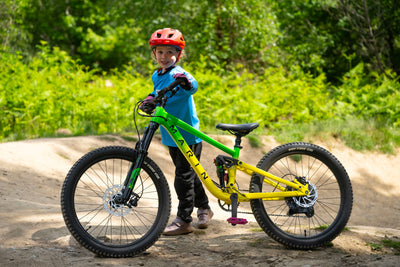 A child wearing a red helmet and blue jacket stands next to a bright green and yellow Arelin full-suspension mountain bike on a dirt trail with green foliage in the background.