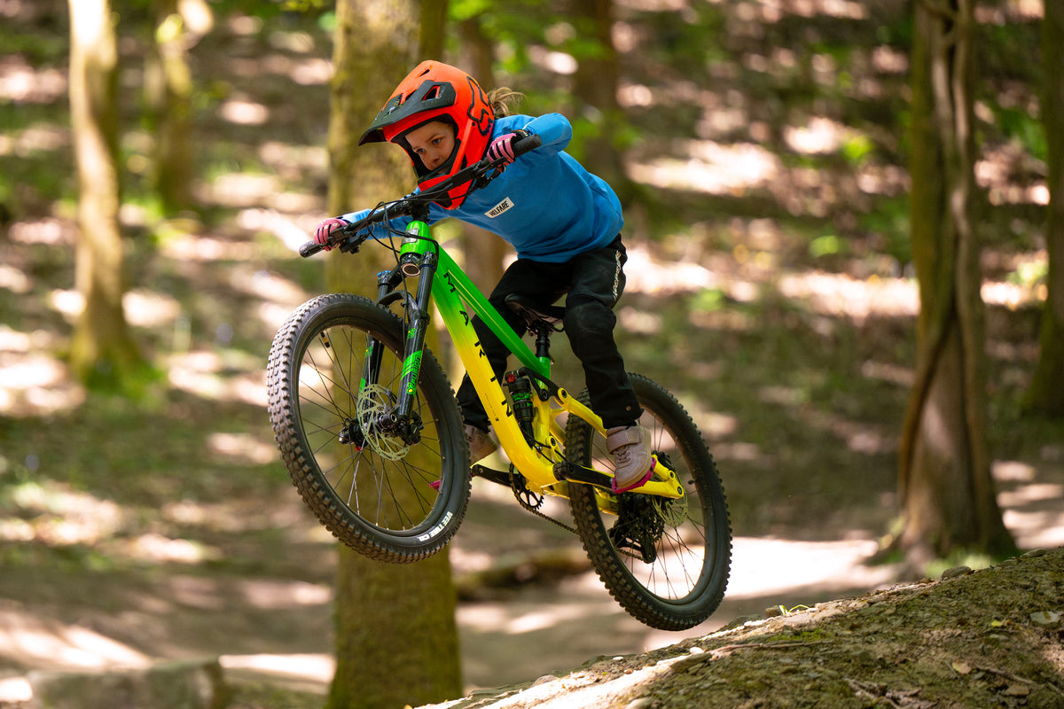A mountain biker wearing a red helmet, blue jacket, and black pants rides a green and yellow full-suspension mountain bike over a dirt trail in a wooded forest setting.