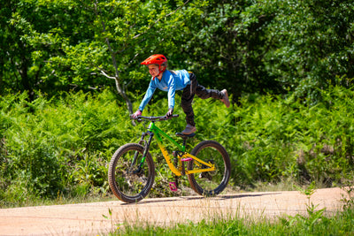 A child wearing a red helmet, blue long-sleeve shirt, and dark pants rides a green and yellow mountain bike on a dirt path surrounded by lush green trees and bushes.