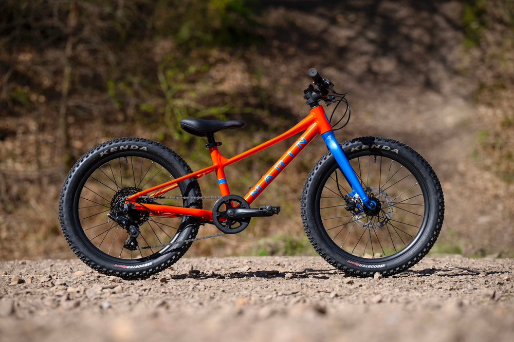 A kids' mountain bike with a bright orange frame, blue front fork, and black tires, shown in profile on a dirt path with a blurred natural background.