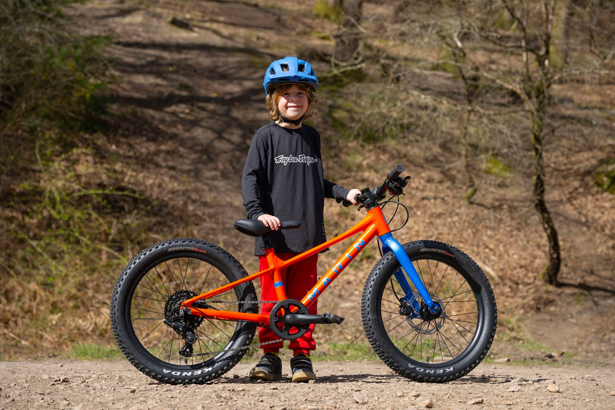 A child wearing a blue helmet, black long-sleeve shirt, and red pants stands next to a fat-tire mountain bike with an orange frame and blue front fork on a dirt path in a wooded outdoor setting.