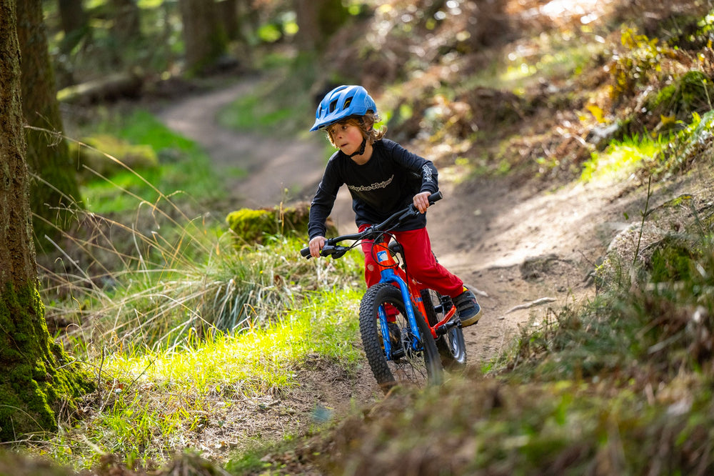 A child wearing a blue helmet, black long-sleeve top, and red pants rides a red and blue mountain bike on a dirt trail in a forest.