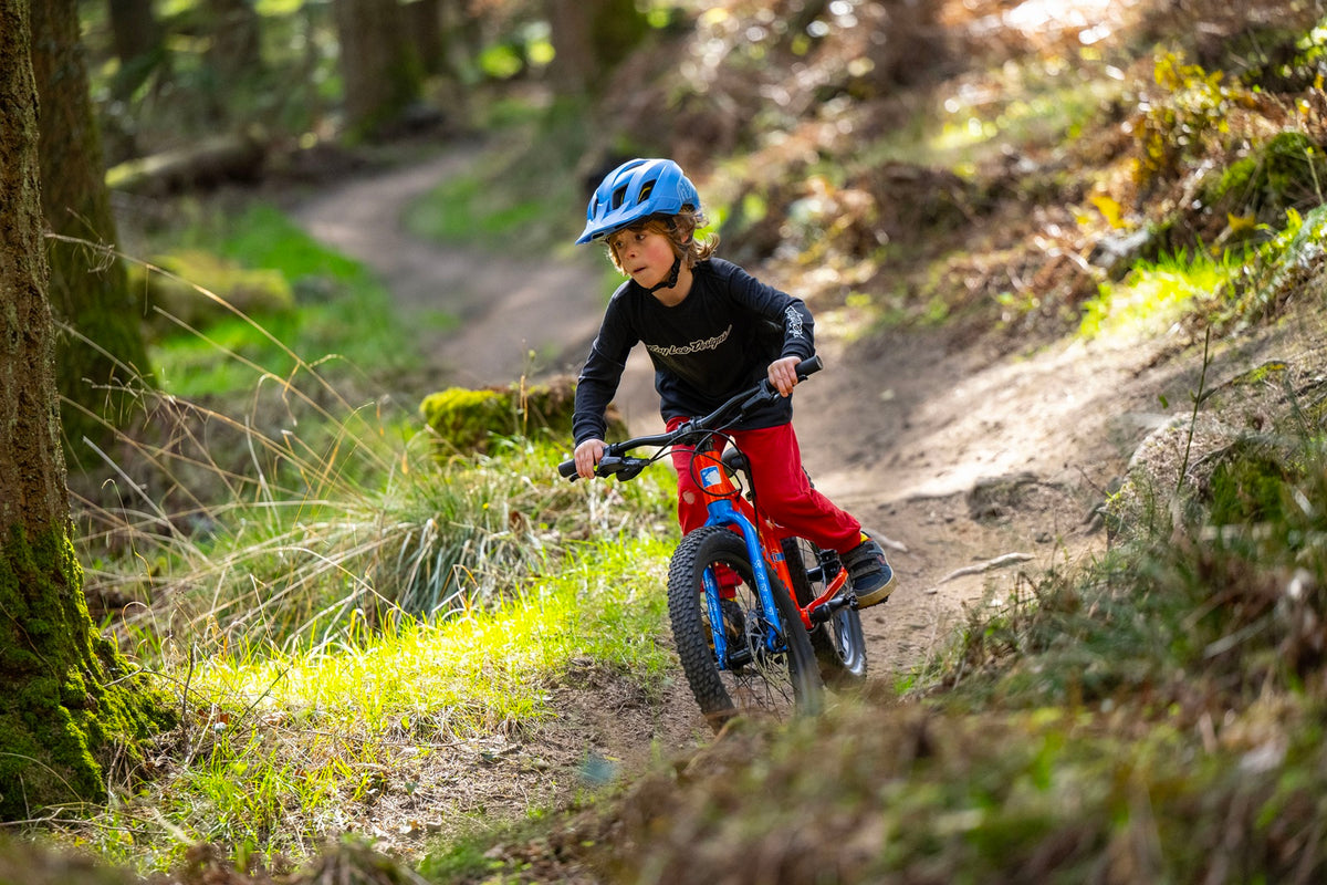 A child wearing a blue helmet, black long-sleeve top, and red pants rides a red and blue mountain bike on a dirt trail in a forest.
