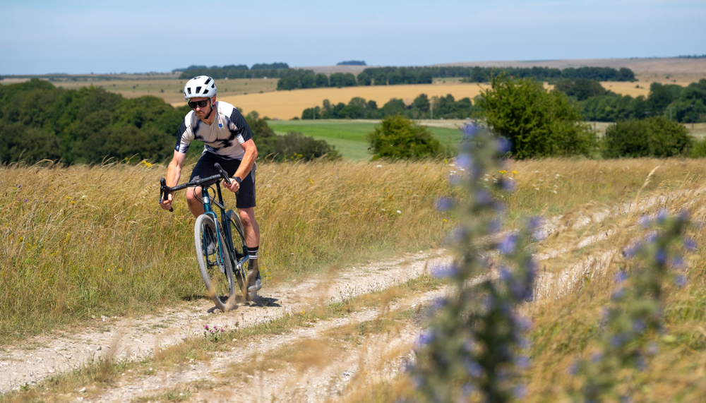 A cyclist rides a gravel bike on a dirt path through a grassy field, with trees and open land in the background under a blue sky.