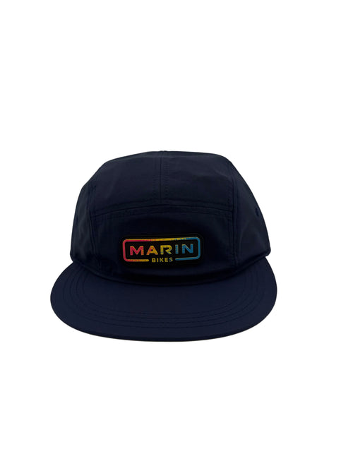 A dark navy blue 5-panel camp cap with a rectangular embroidered front patch reading 'MARIN DIES' in a rainbow gradient, set against a neutral light gray background.