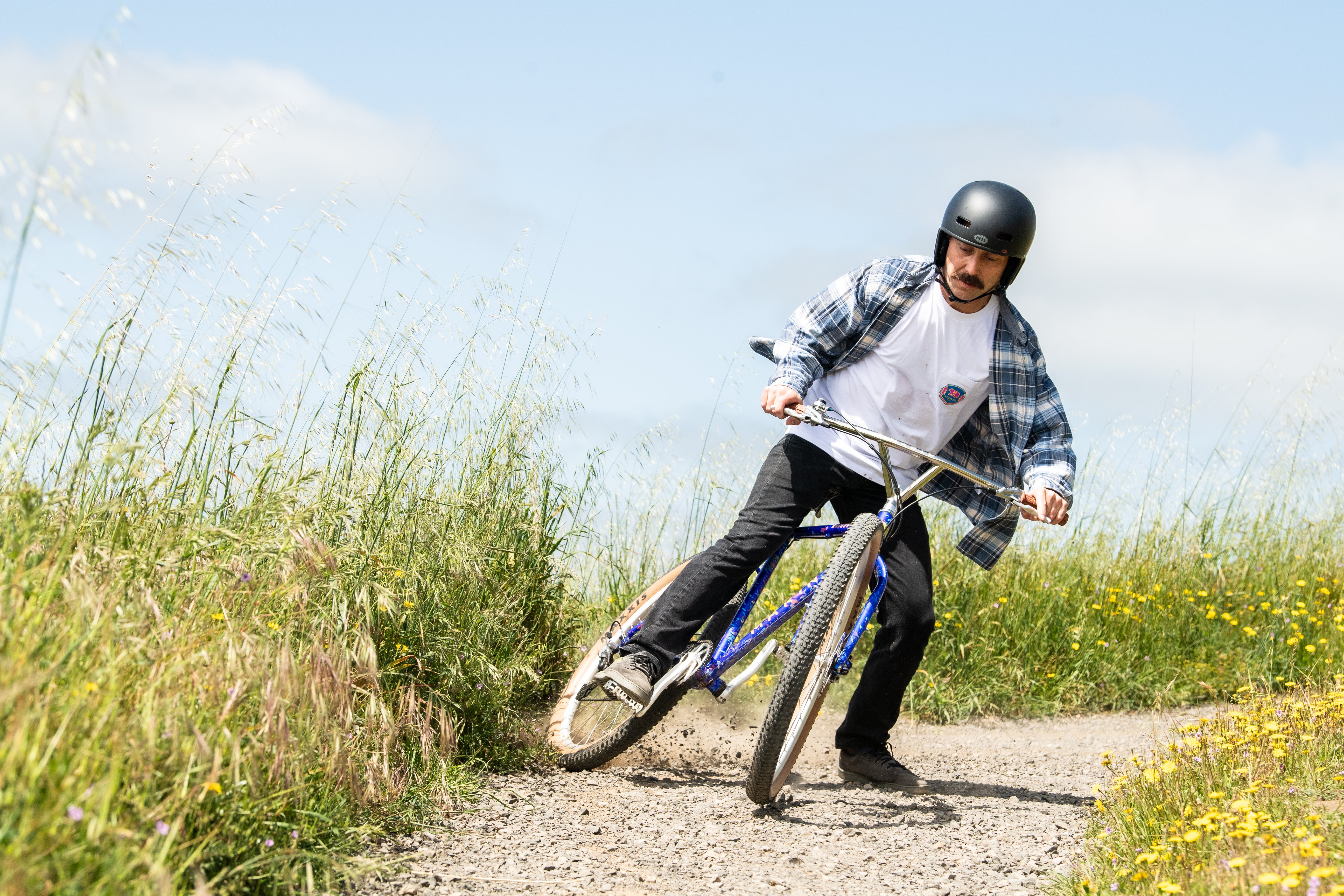 A man wearing a black helmet rides a blue mountain bike on a gravel path surrounded by tall grass and yellow wildflowers under a blue sky.
