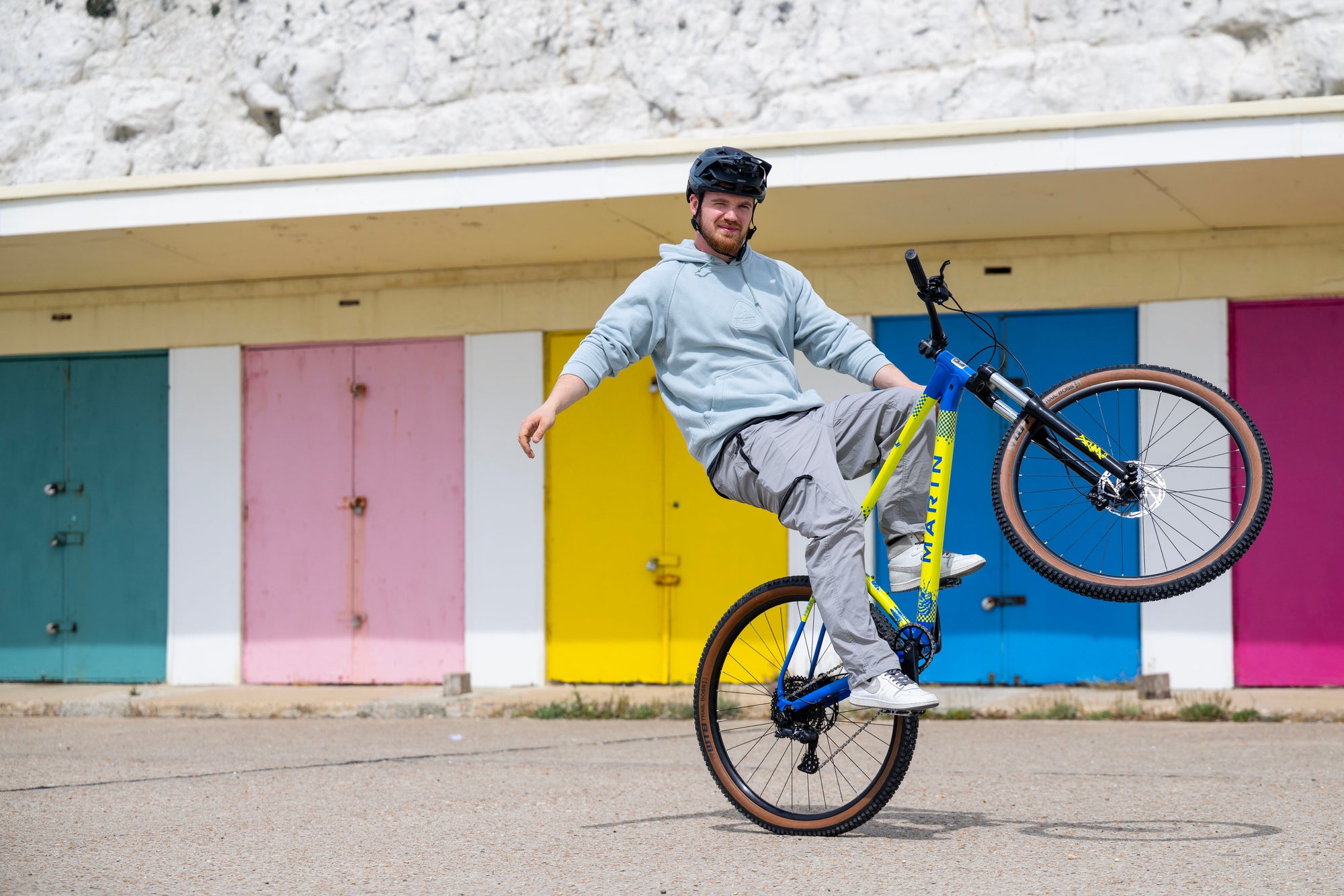 A man wearing a black helmet, light blue hoodie, and gray sweatpants performs a wheelie on a yellow and blue mountain bike in front of a building with colorful garage-style doors.