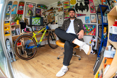 A man sits in an office chair in a cluttered, highly decorated room with a yellow and blue mountain bike behind him. The room is filled with posters, skateboards, shelves, and collectibles, creating a busy, creative atmosphere.