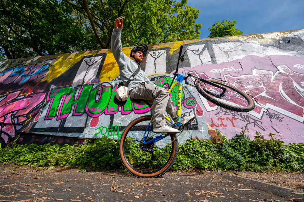 A person riding a blue and yellow mountain bike while balancing on one wheel in front of a graffiti-covered concrete wall outdoors.