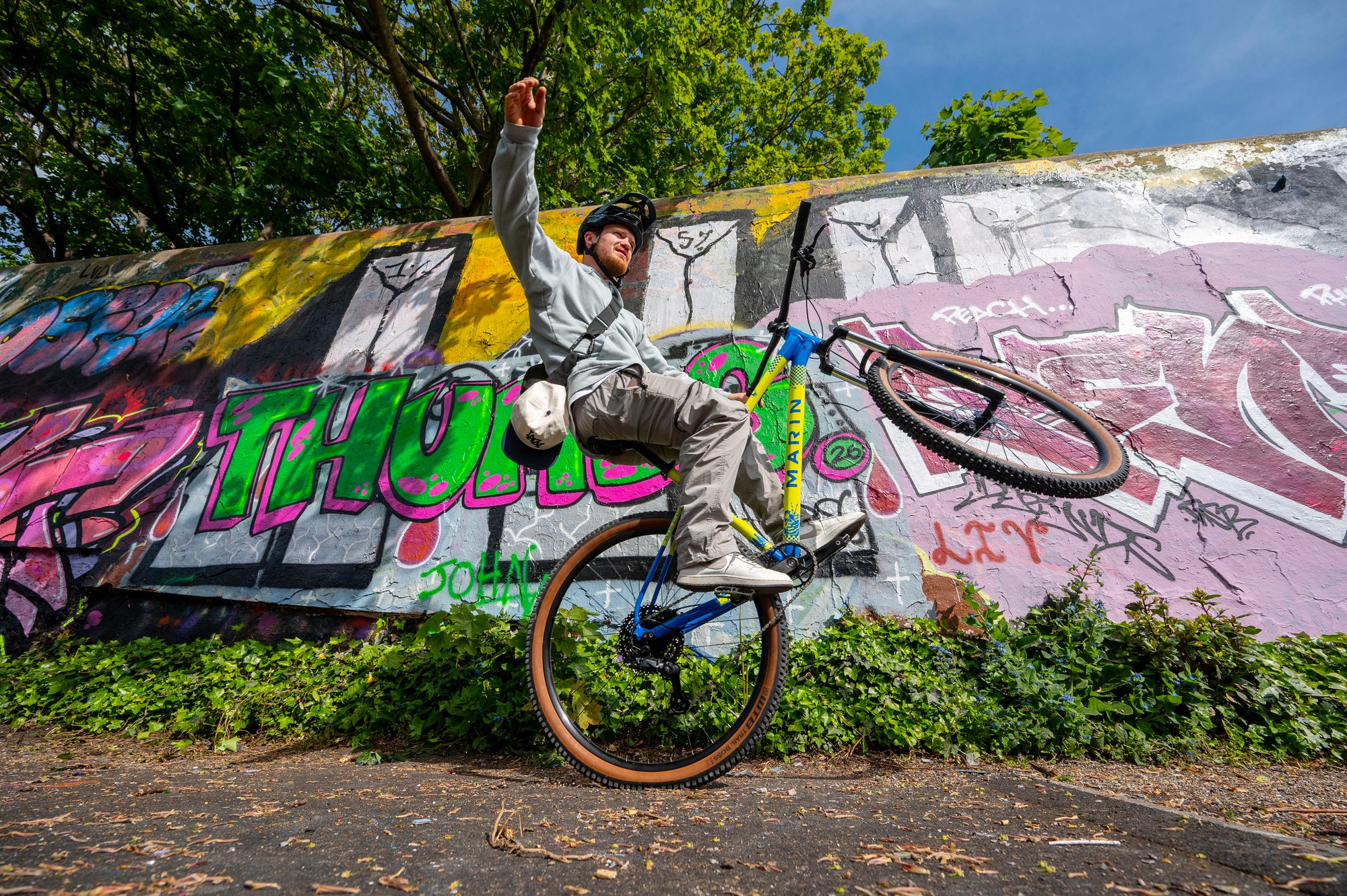 A person riding a blue and yellow mountain bike while balancing on one wheel in front of a graffiti-covered concrete wall outdoors.