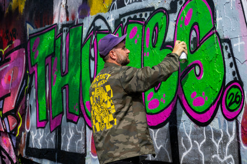 A man wearing a purple cap and a camouflage jacket with a yellow graphic on the back, spray painting large green and pink graffiti letters on a gray concrete wall.
