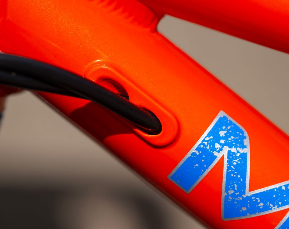 Close-up of a bright orange bicycle frame with a blue and white stylized 'M' logo and a black cable running through a cable housing.