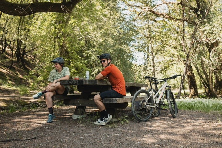 Two cyclists sitting at a picnic table in a forest with their bikes nearby.