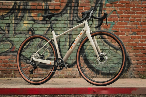 White bicycle with brown tires leaning against a brick wall with graffiti.