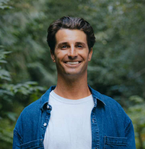 Man wearing a blue denim jacket over a white shirt with a blurred green background