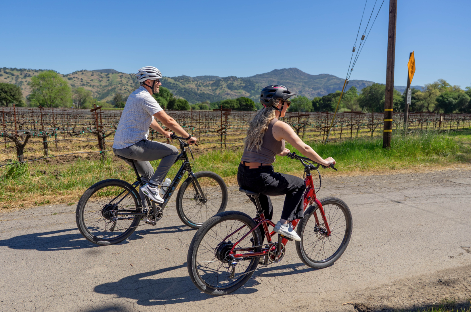 Two people riding bicycles on a road with a scenic background of mountains and trees.