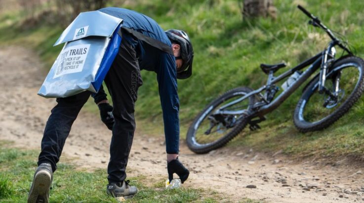 An image of a male mountain biker picking up litter from the trail with a Marin Bikes mountain bike placed at the side of the trail